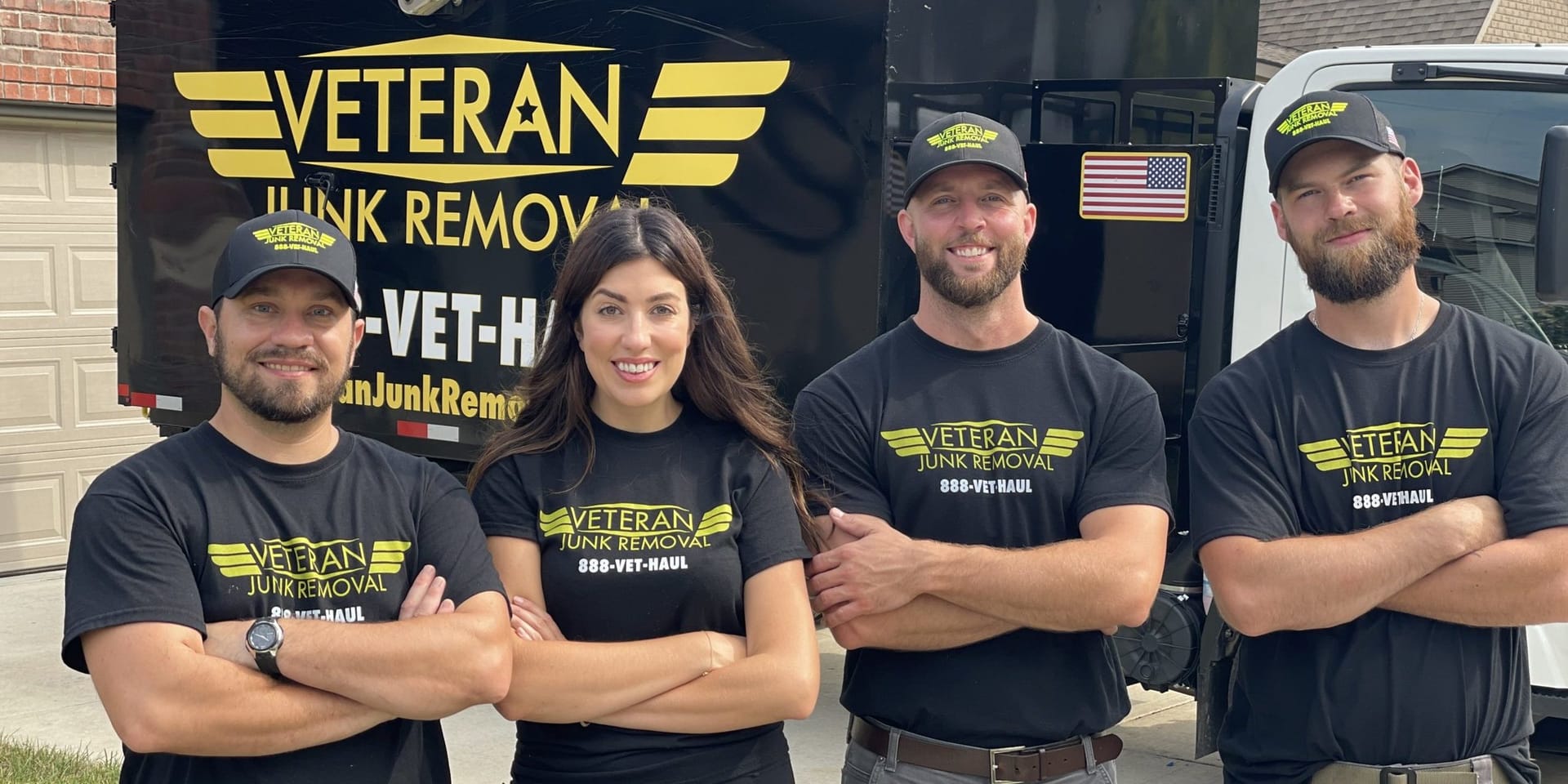 Four Veteran Junk Removal team members with arms crossed in front of the truck's winged logo and a US flag decal