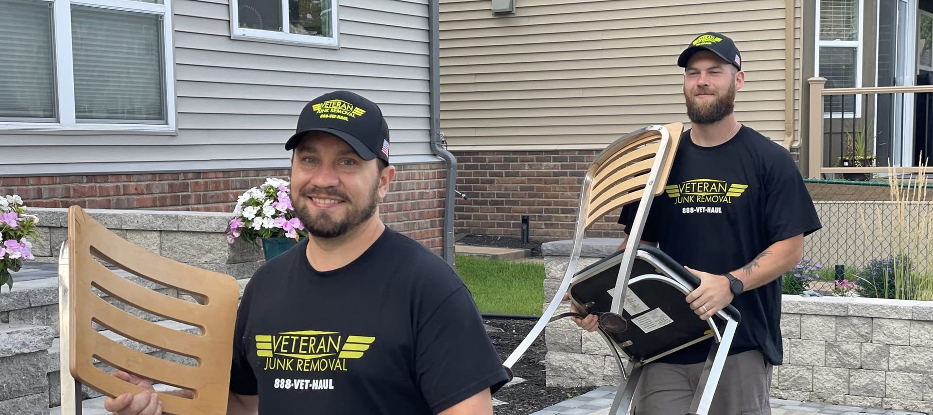 Two Veteran Junk Removal crew members carrying patio chairs out from a home's backyard patio