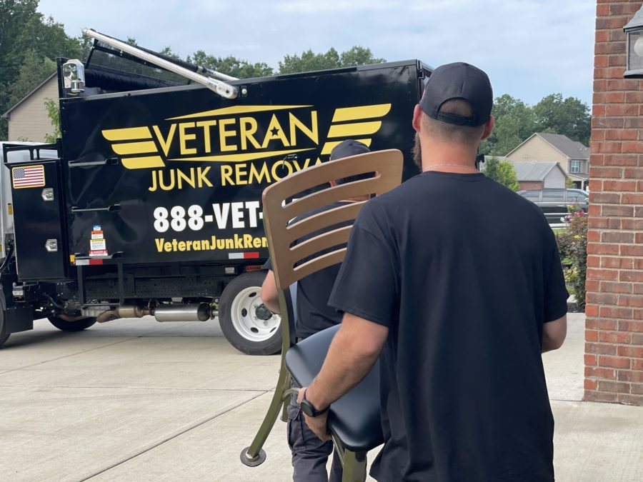 Veteran Junk Removal crew carrying a chair down a driveway toward the branded truck