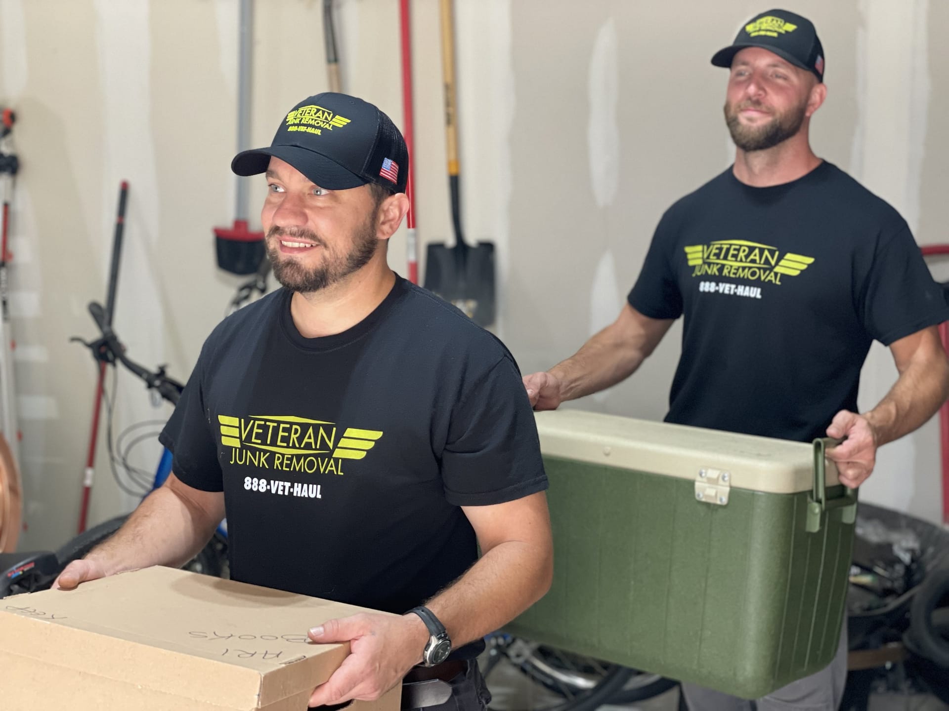 Two Veteran Junk Removal crew members carrying a box and a cooler out of a garage