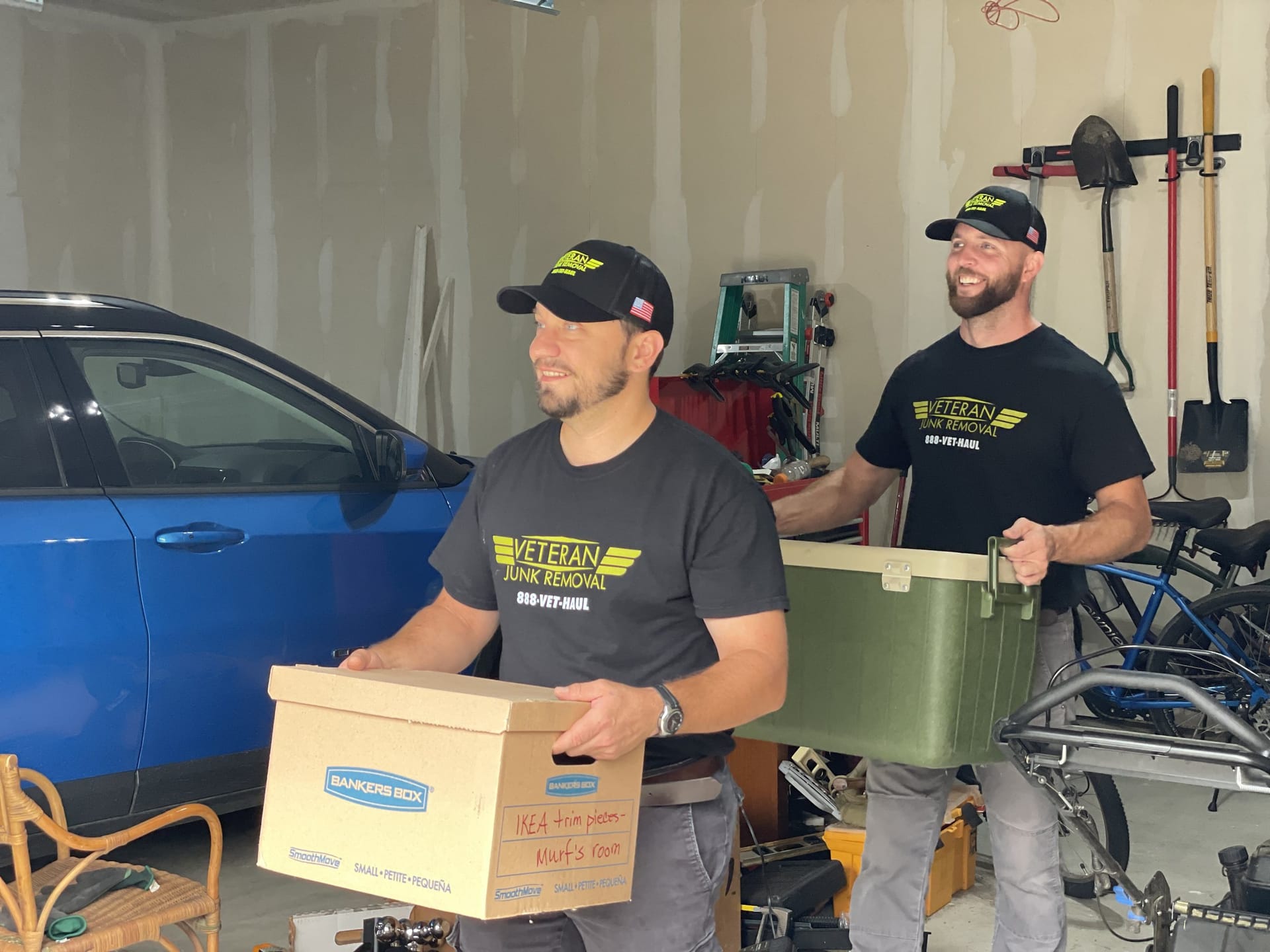 Two Veteran Junk Removal crew members carrying a storage box and a cooler through a garage past a parked car